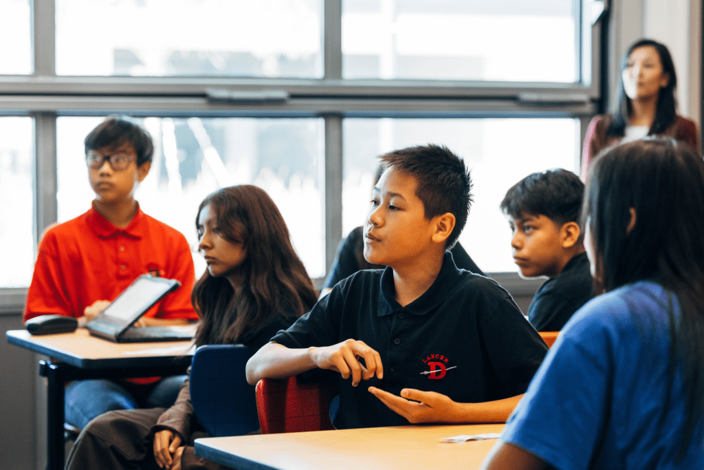 Middle school students seated at desks during class, with one student speaking while others listen attentively. A teacher stands in the background near the window.