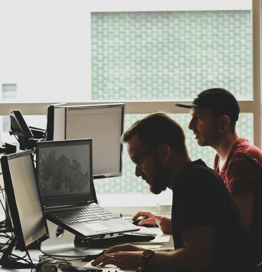 Two people working at a desk with multiple computer monitors in a modern, naturally lit workspace.