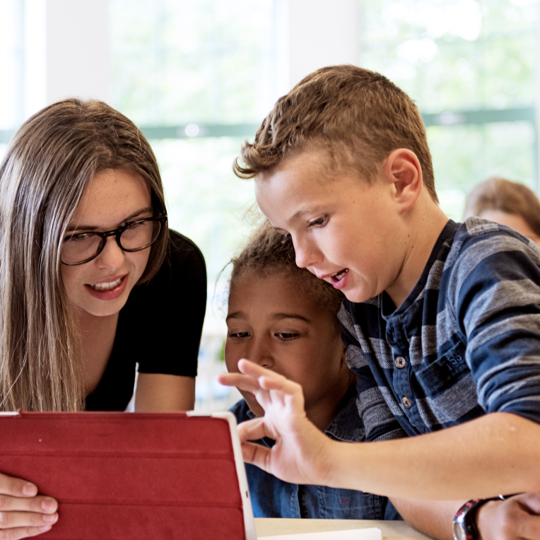 Two children and an adult gather around a tablet, engaged and smiling as they interact with the screen in a bright classroom setting.