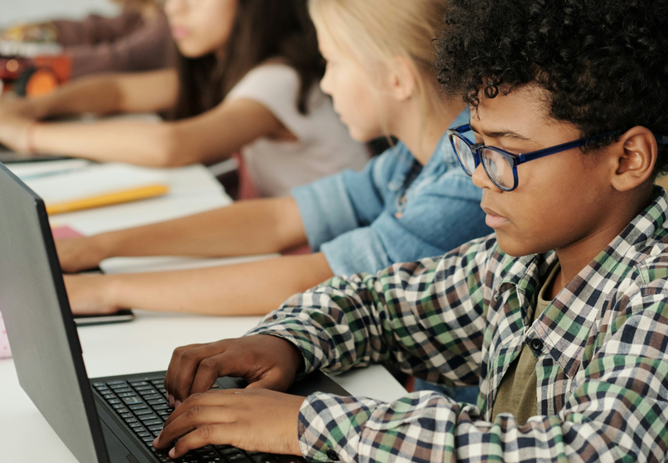 Children sitting at a table working on laptops, focused on their tasks in a classroom or tech lab setting.