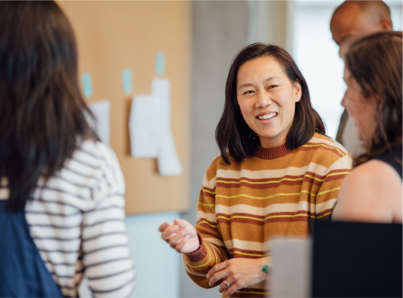 Priscilla Chan in a striped sweater smiling and talking with colleagues in a collaborative workspace, with notes pinned to a board in the background.