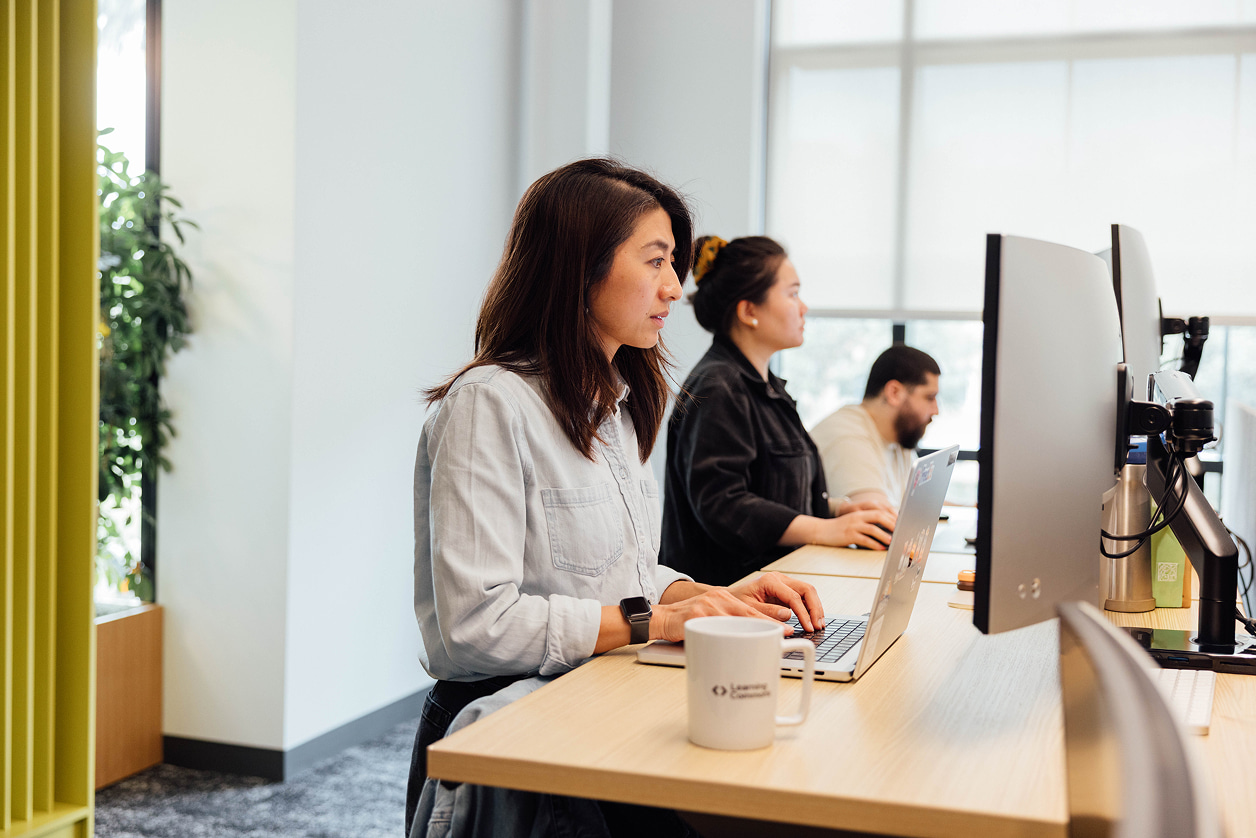 Three people, two standing and one sitting, working side by side on computers at an elongated shared workspace