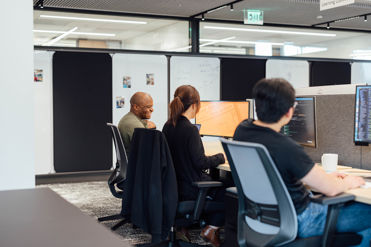 Three people seated and working side by side on computers at an elongated shared workspace