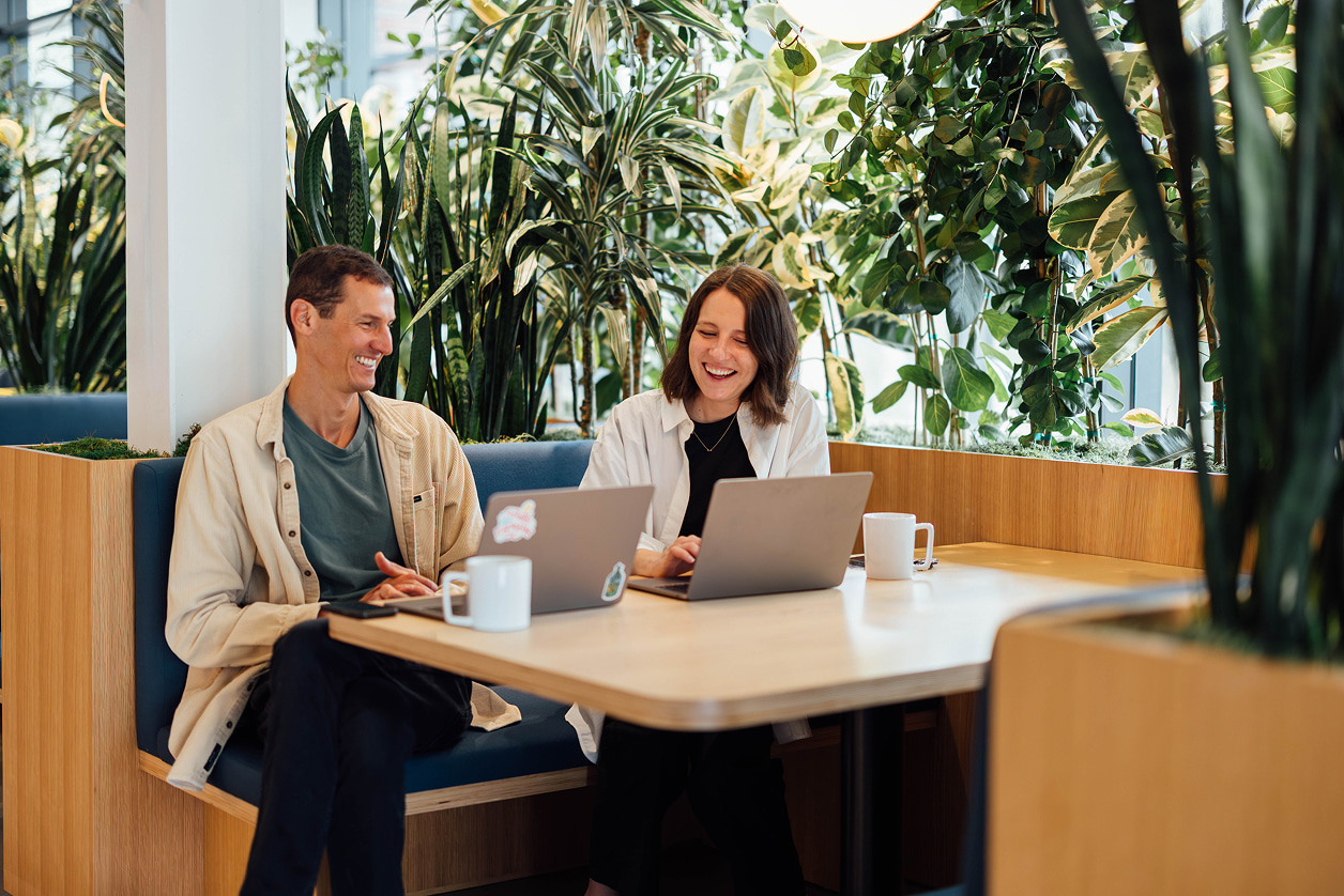 Two people smiling and working on laptops together in a plant-filled office lounge booth