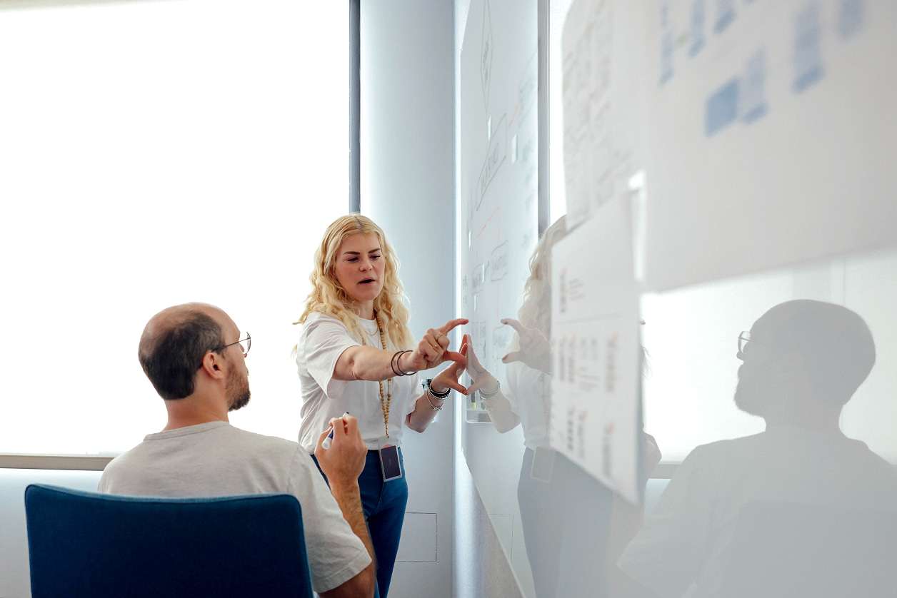 Two people in a bright meeting room discussing content on a whiteboard covered in notes