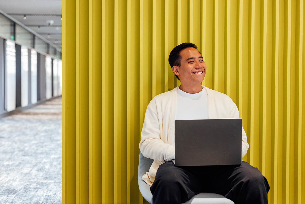 A person sitting in a chair with a laptop, smiling, against a yellow ribbed wall