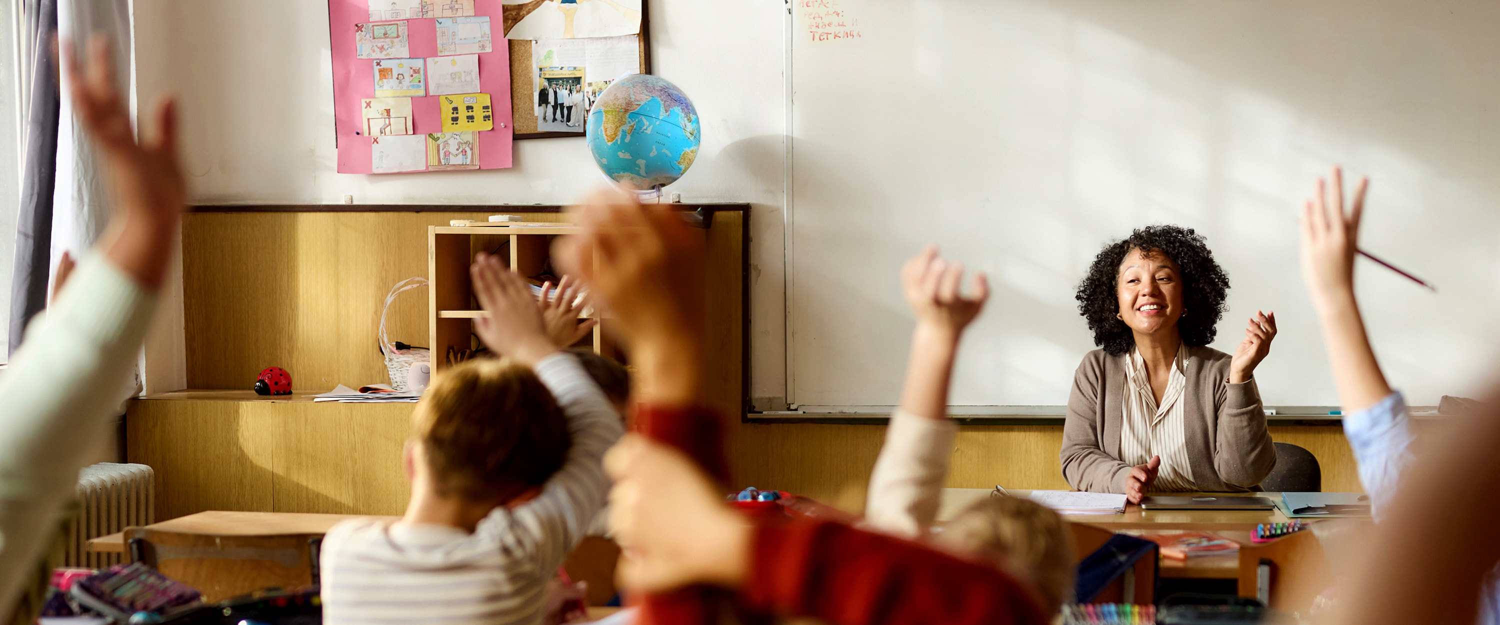 Smiling teacher at her desk engages an enthusiastic class of students with raised hands