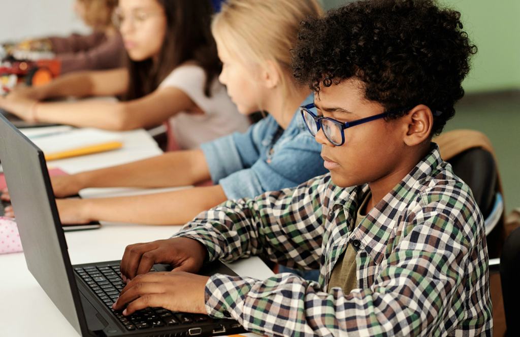 A young student wearing glasses working on a laptop in a classroom with other students in the background