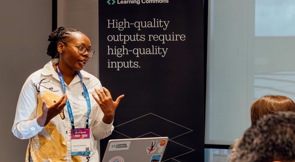A presenter speaking in front of a Learning Commons banner reading "High-quality outputs require high-quality inputs."